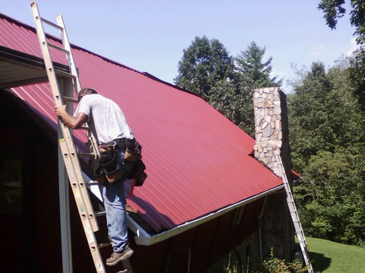 Roofer on a ladder beside a red metal roof and stone chimney — Soffit & Fascia Repair by Roofing Brothers in Villa Rica